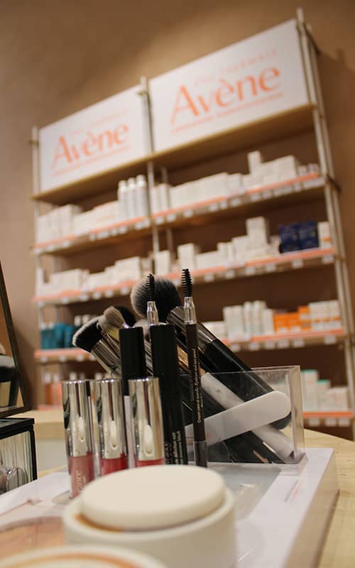 Gros plan sur des pinceaux de maquillage et des gloss disposés sur un présentoir, avec des étagères de produits de la marque Avène en arrière-plan flou.