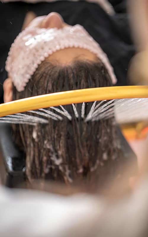 Gros plan sur un soin capillaire relaxant : un anneau doré diffuse de fins jets d'eau sur les cheveux d'une personne allongée.