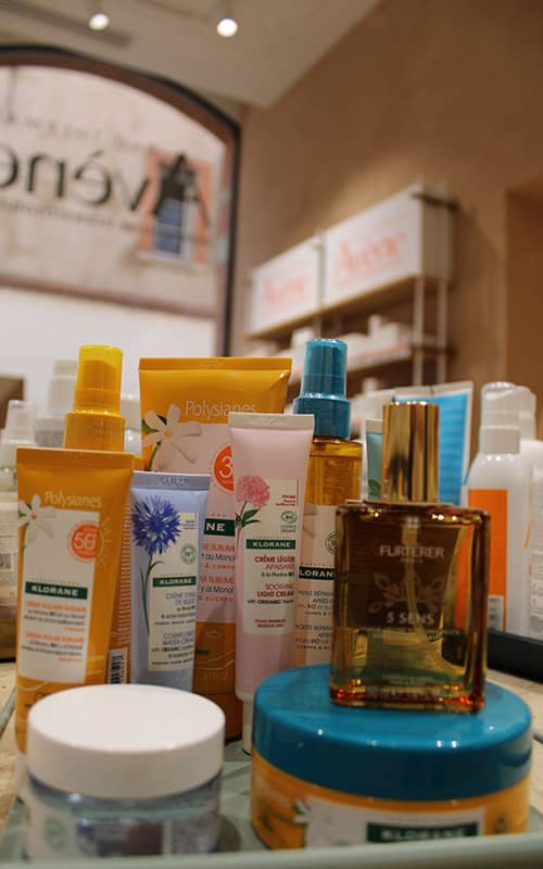 Gros plan sur une sélection de produits de soin : crèmes solaires Polysianes, tubes de crème Klorane (au bleuet et à la pivoine) et une bouteille d'huile sèche '5 Sens' de René Furterer. En arrière-plan flou, on distingue des rayons de pharmacie avec l'enseigne Avène.
