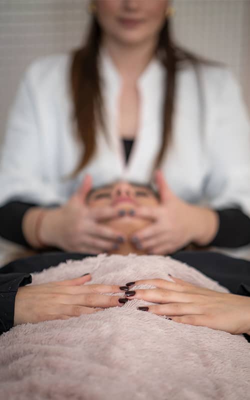 Vue en contre-plongée d'une séance de bien-être. Au premier plan, les mains de la cliente reposent sur une couverture en fausse fourrure rose. En arrière-plan, une professionnelle masse délicatement le visage de la cliente.